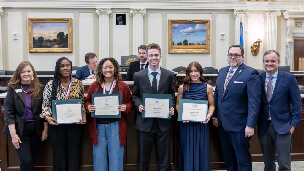 Group of students and officials stand in a government chamber holding certificates and awards during a recognition ceremony.