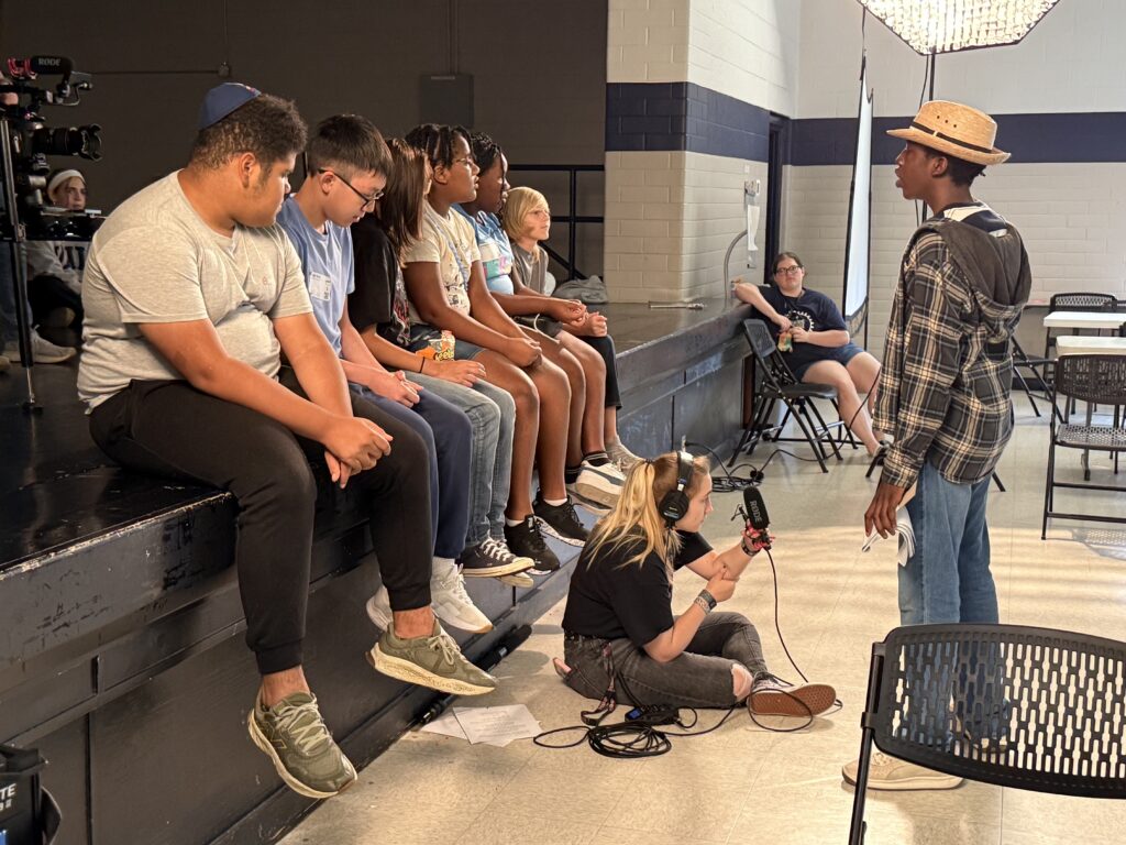 Students participate in a film production exercise, with one student acting on stage while others observe and a crew member records audio with a microphone.