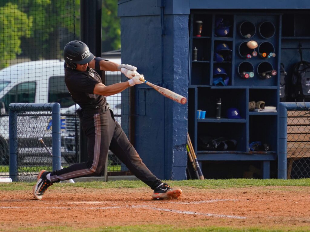 Rose State College baseball player swings at a pitch during a game, with the dugout visible in the background.