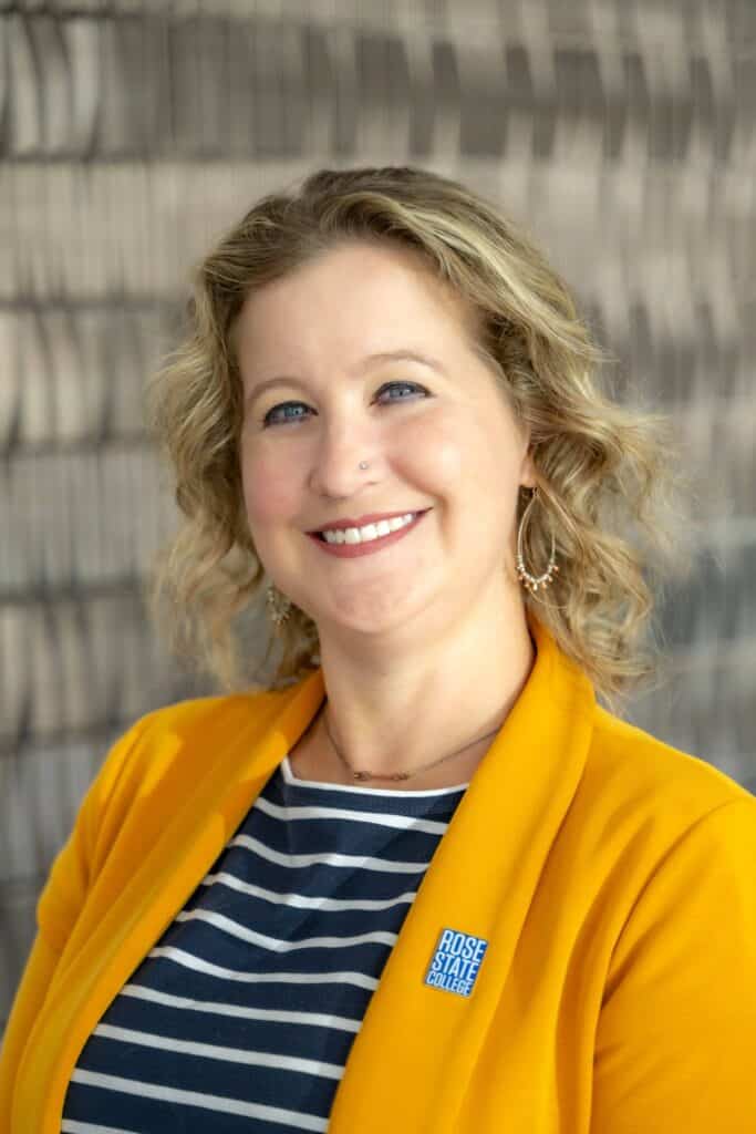 Professional headshot of a smiling woman with curly blonde hair, wearing a mustard yellow blazer and a navy striped shirt with a Rose State College pin, in front of a softly blurred neutral background.