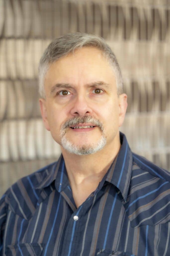 Professional headshot of a man with short gray hair and a goatee, wearing a dark blue striped shirt, in front of a softly blurred neutral background.