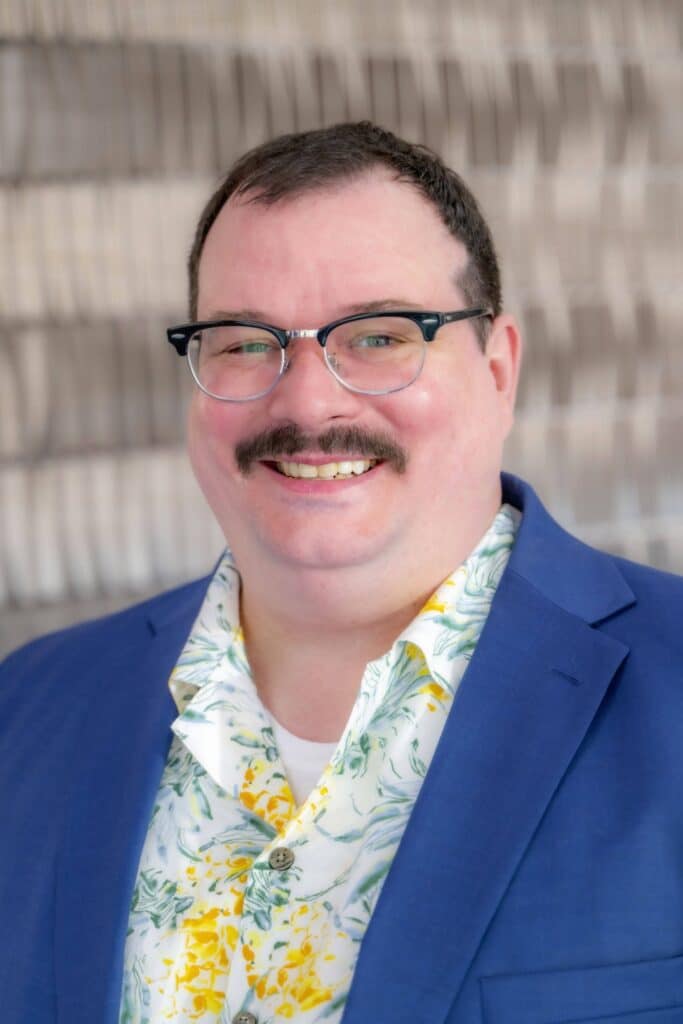Professional headshot of a smiling man with short dark hair, glasses, and a mustache, wearing a blue blazer and a floral shirt, in front of a softly blurred neutral background."