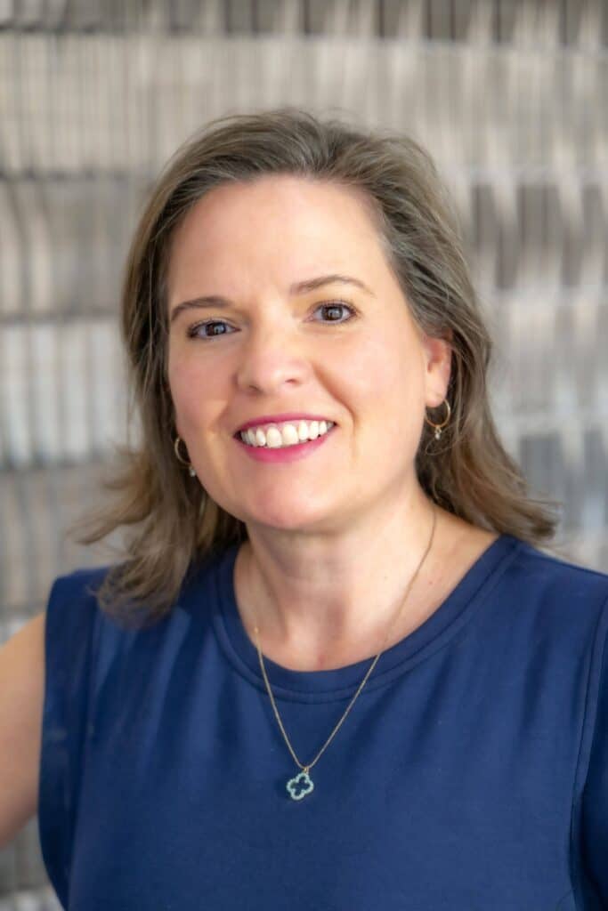 Professional headshot of a smiling woman with shoulder-length brown hair, wearing a navy blue top and a clover-shaped necklace, in front of a softly blurred neutral background.