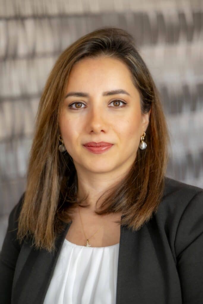 Professional headshot of a woman with straight brown hair, wearing a black blazer and white blouse, in front of a softly blurred neutral background.