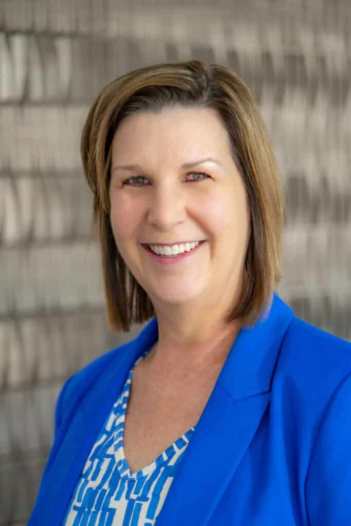 Professional headshot of a smiling woman with short brown hair, wearing a royal blue blazer and patterned blue and white blouse, in front of a softly blurred neutral background.