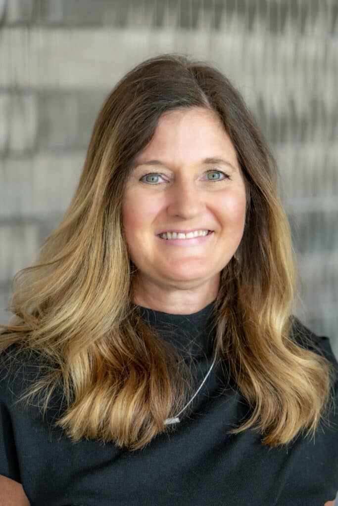 Professional headshot of a smiling woman with long light brown hair, wearing a black top and silver necklace, in front of a softly blurred neutral background.