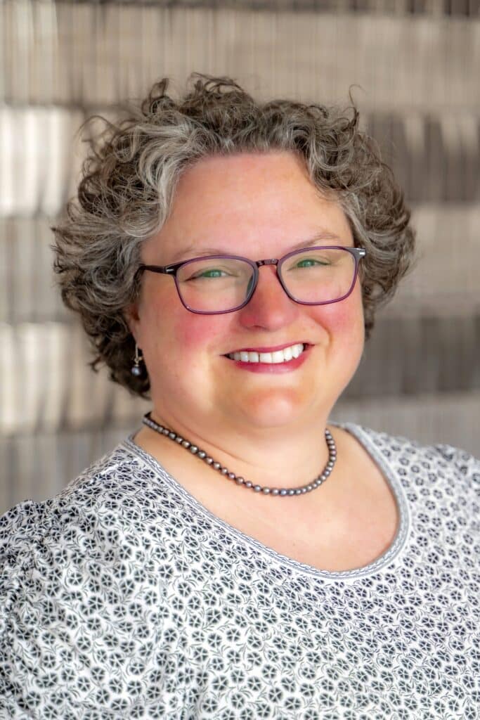 Professional headshot of a smiling woman with short curly gray hair and glasses, wearing a patterned white and black top and a pearl necklace, in front of a softly blurred neutral background.
