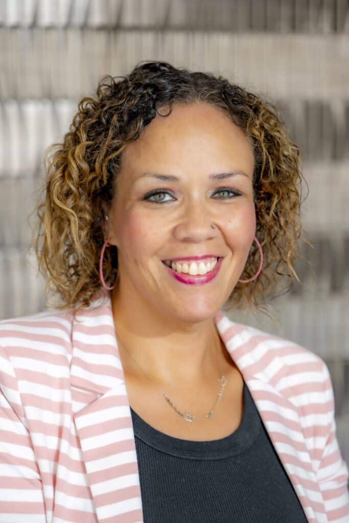 Professional headshot of a smiling man with glasses and a short beard wearing a light blue button-up shirt, in front of a softly blurred neutral background.