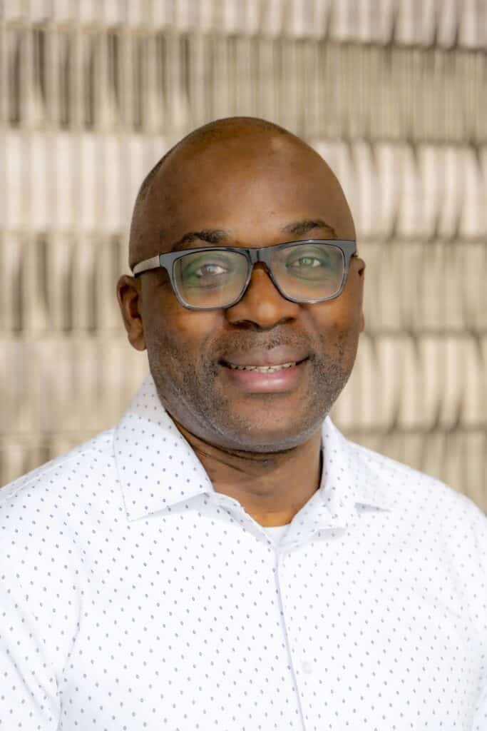 Professional headshot of a smiling man wearing glasses and a white dress shirt with a subtle pattern, in front of a softly blurred neutral background.