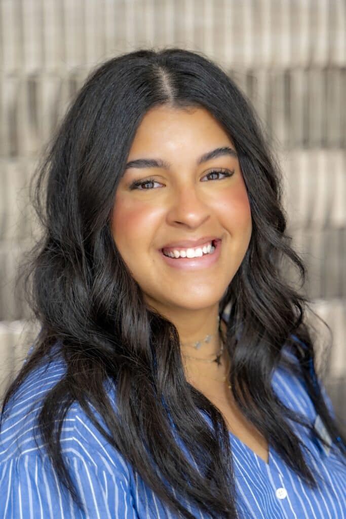 Professional headshot of a smiling woman with long dark hair wearing a blue and white striped blouse, in front of a softly blurred neutral background.