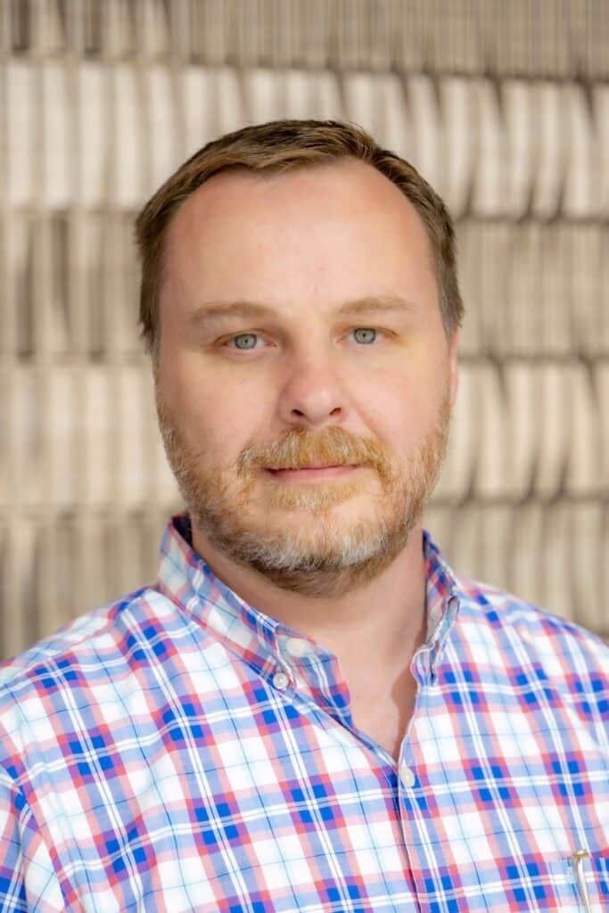 Professional headshot of a man with short hair and a beard wearing a red, white, and blue plaid shirt, in front of a softly blurred neutral background.