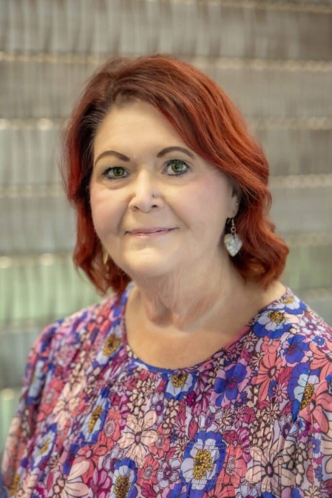 Professional headshot of a woman with short red hair wearing a colorful floral blouse and heart-shaped earrings, in front of a softly blurred neutral background.