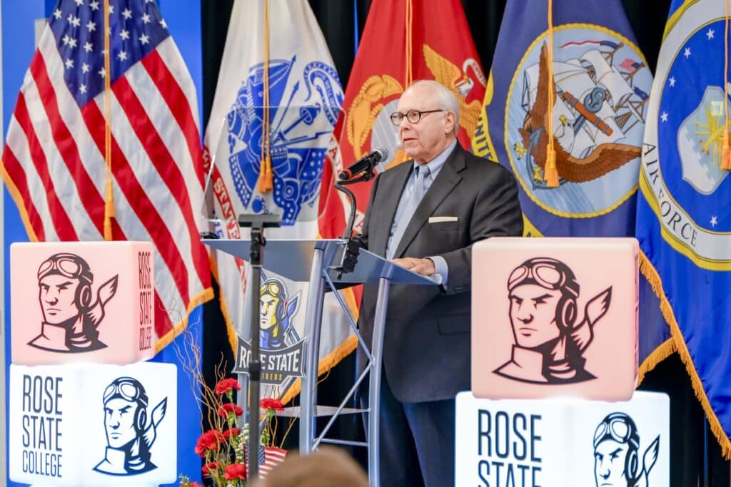 An older man in a suit speaks at a podium during a Rose State College Veterans Day event, with U.S. military branch flags and Rose State College signage displayed behind him.