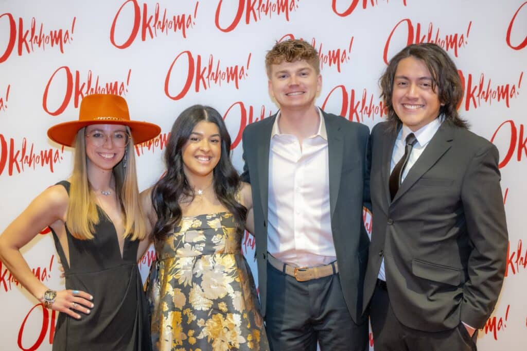 A group of four young adults dressed in formal attire smiling in front of a step-and-repeat backdrop that reads 'Oklahoma!' in red lettering.