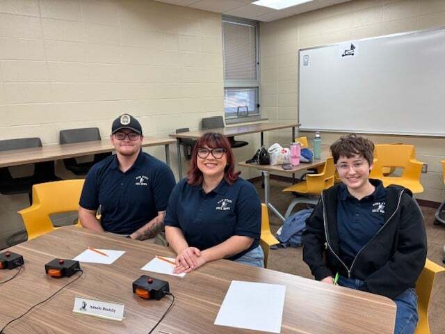 Three students wearing matching navy shirts sit at a table with quiz buzzers and paper in a classroom.