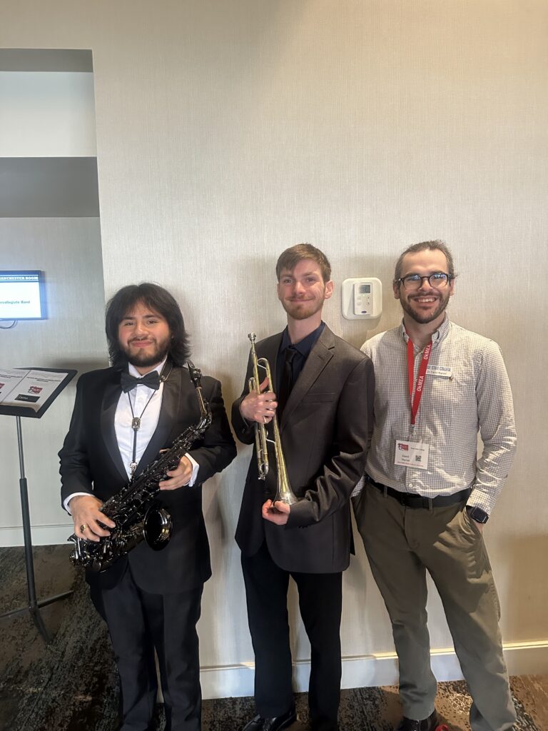 Two student musicians in formal concert attire stand holding a saxophone and a trumpet beside a Rose State College faculty member wearing a name badge, indoors at a music event.