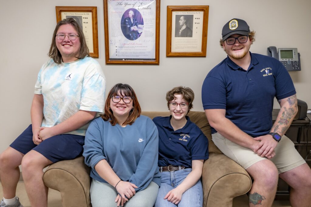 Four Rose State College Quiz Bowl team members sit and stand together on a couch, smiling for a group photo in a campus office.