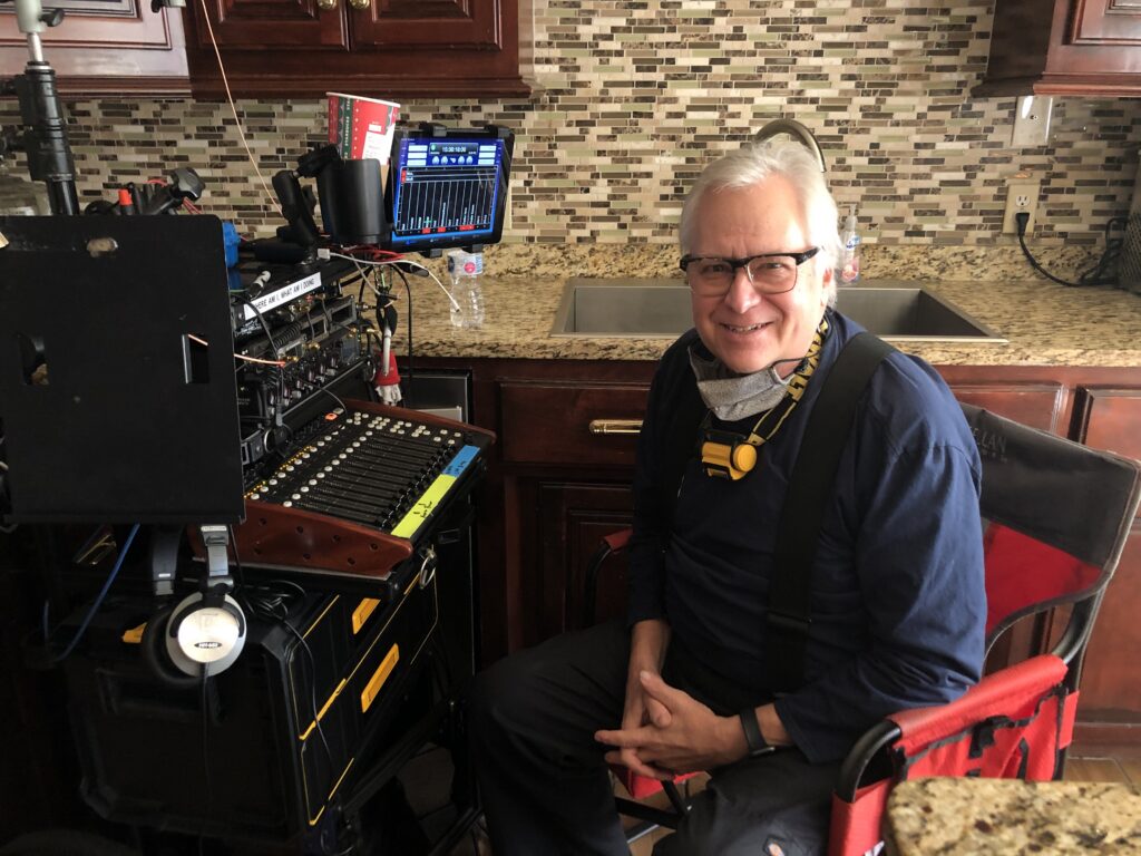 **Alt text:** A man wearing glasses sits beside an audio mixing board and recording equipment set up on a cart in a kitchen area.