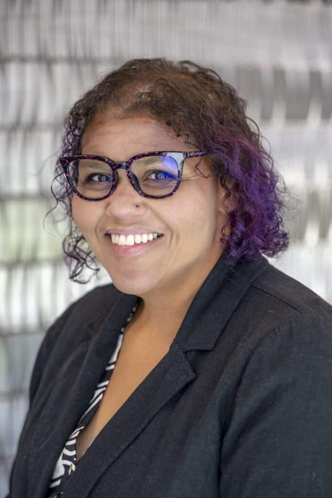 Professional headshot of a smiling woman wearing glasses and a black blazer, with curly brown and purple hair, standing in front of a light textured background.