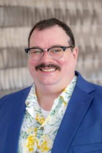 Professional headshot of a smiling man with short dark hair, glasses, and a mustache, wearing a blue blazer and a floral shirt, in front of a softly blurred neutral background."