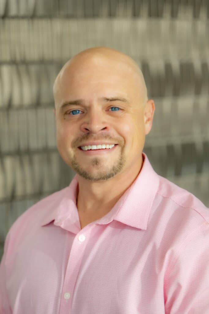 Professional headshot of a smiling man with a shaved head and goatee, wearing a light pink collared shirt, in front of a softly blurred neutral background.