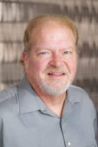 Professional headshot of a smiling man with short light hair and a trimmed beard, wearing a gray collared shirt, in front of a softly blurred neutral background.