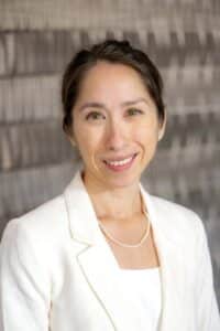 Professional headshot of a smiling woman with dark hair pulled back, wearing a white blazer and pearl necklace, in front of a softly blurred neutral background.