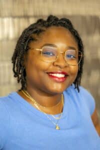 Professional headshot of a smiling woman with short braided hair wearing gold glasses, a blue top, and gold necklaces, in front of a softly blurred neutral background.