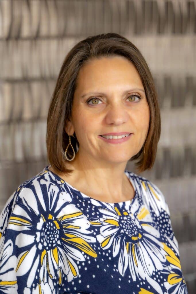 Professional headshot of a woman with short brown hair wearing a navy and white floral top with yellow accents, smiling in front of a softly blurred neutral background.