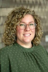 Professional headshot of a smiling woman with curly blonde hair and glasses wearing a green blouse with a subtle dotted pattern, in front of a softly blurred neutral background.