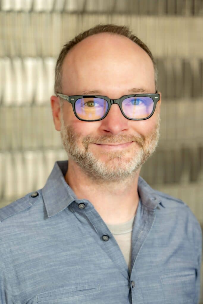 Professional headshot of a smiling man with glasses and a short beard wearing a light blue button-up shirt, in front of a softly blurred neutral background.