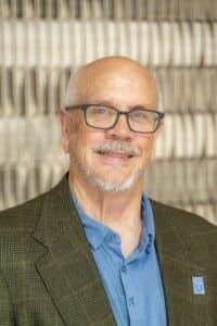 Professional headshot of a smiling man with glasses and a short gray beard wearing a blue shirt and green plaid blazer with a Rose State College pin, in front of a softly blurred neutral background.