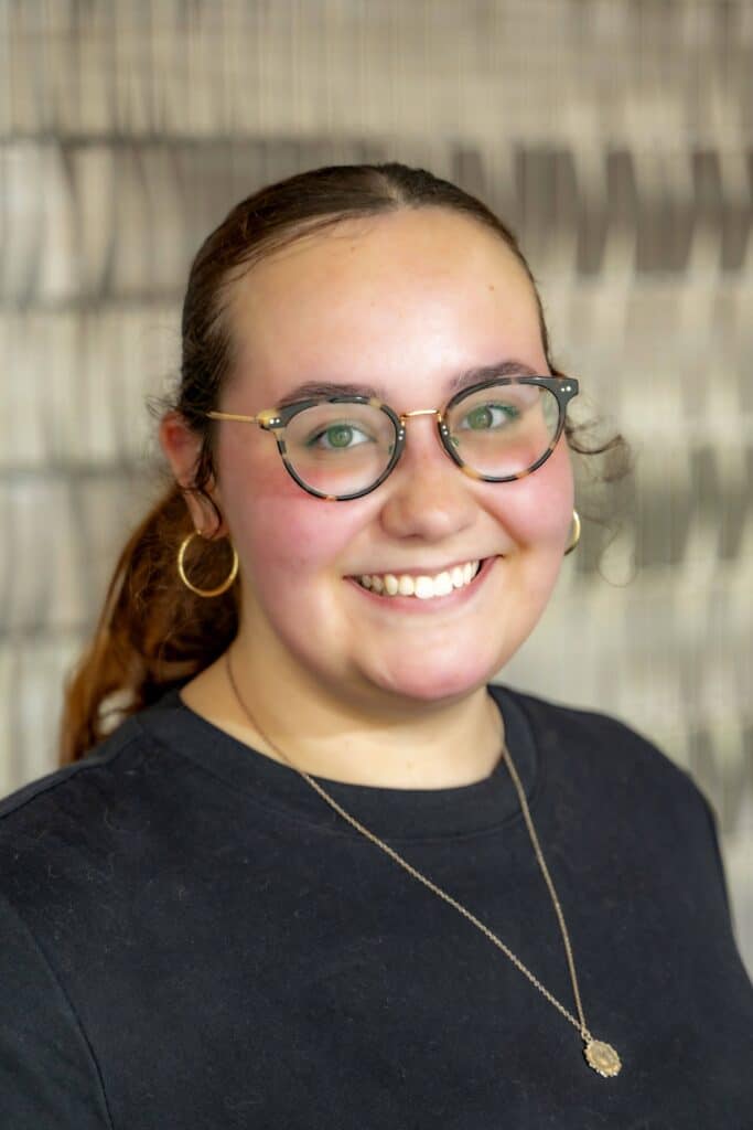 Professional headshot of a smiling woman with glasses and pulled-back hair wearing a black top and gold necklace, in front of a softly blurred neutral background.