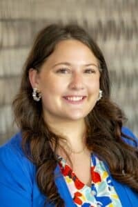 Professional headshot of a smiling woman with long brown hair, wearing pearl earrings, a blue blazer, and a colorful floral blouse, in front of a softly blurred neutral background.