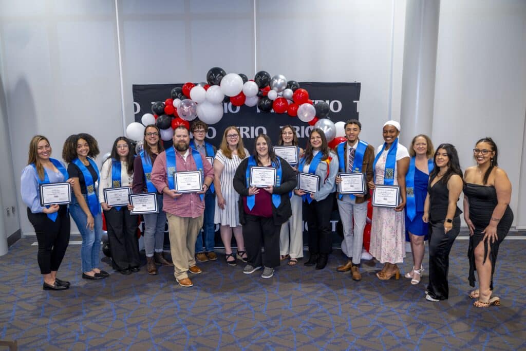 A group of thirteen people stand in a row indoors, smiling at the camera. Most are wearing blue graduation stoles with “Rose State College” printed on them and holding certificates. Behind them is a black backdrop with the word “TRIO” repeated, along with a balloon arch in black, white, red, and silver. The group includes a mix of men and women of varying ages and ethnicities, dressed in business casual and semi-formal attire. The floor is carpeted in blue and gray, and the room has white walls.