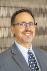 Professional headshot of a man with glasses and a beard, wearing a blue Rose State College polo shirt, in front of a softly blurred neutral background.