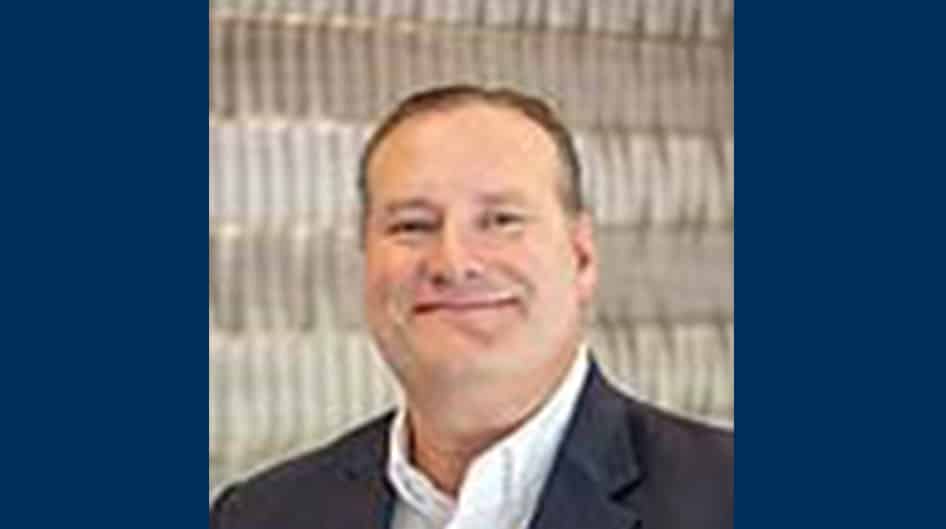 Professional headshot of a smiling man wearing glasses, a dark suit jacket, white dress shirt, and patterned tie, posed in front of a plain dark background.