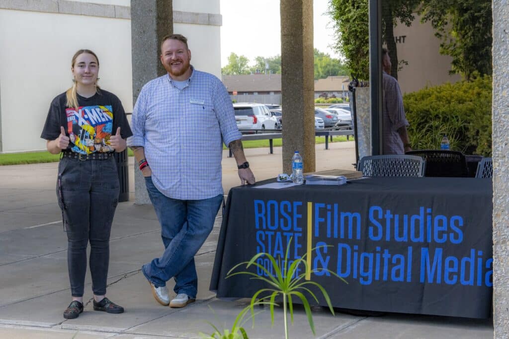 Two people standing and smiling next to a table covered with a Rose State College Film Studies and Digital Media tablecloth. One person is giving two thumbs up, and the other is leaning against the table outside a campus building.