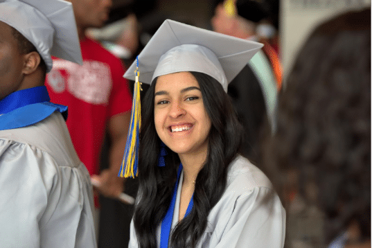 Young woman in a gray graduation cap and gown smiles while seated among other graduates during a commencement ceremony.