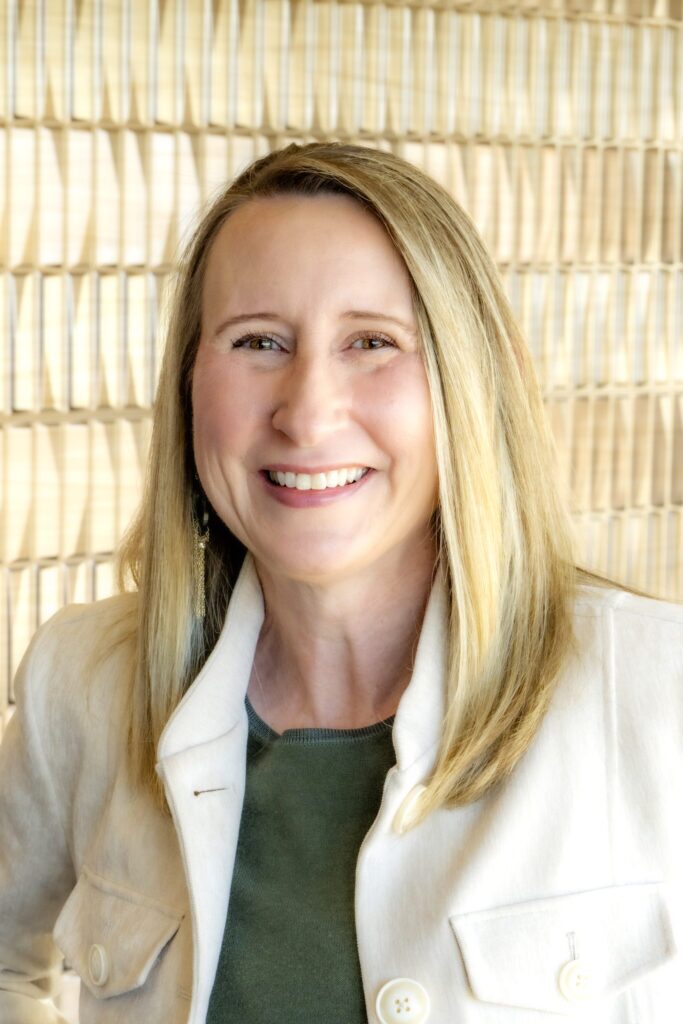 Portrait of a smiling woman with blonde hair wearing a cream jacket over a green top, photographed against a light textured background.