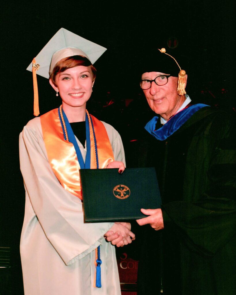 Graduate in a gray cap and gown shakes hands with a faculty member while holding a diploma cover onstage.