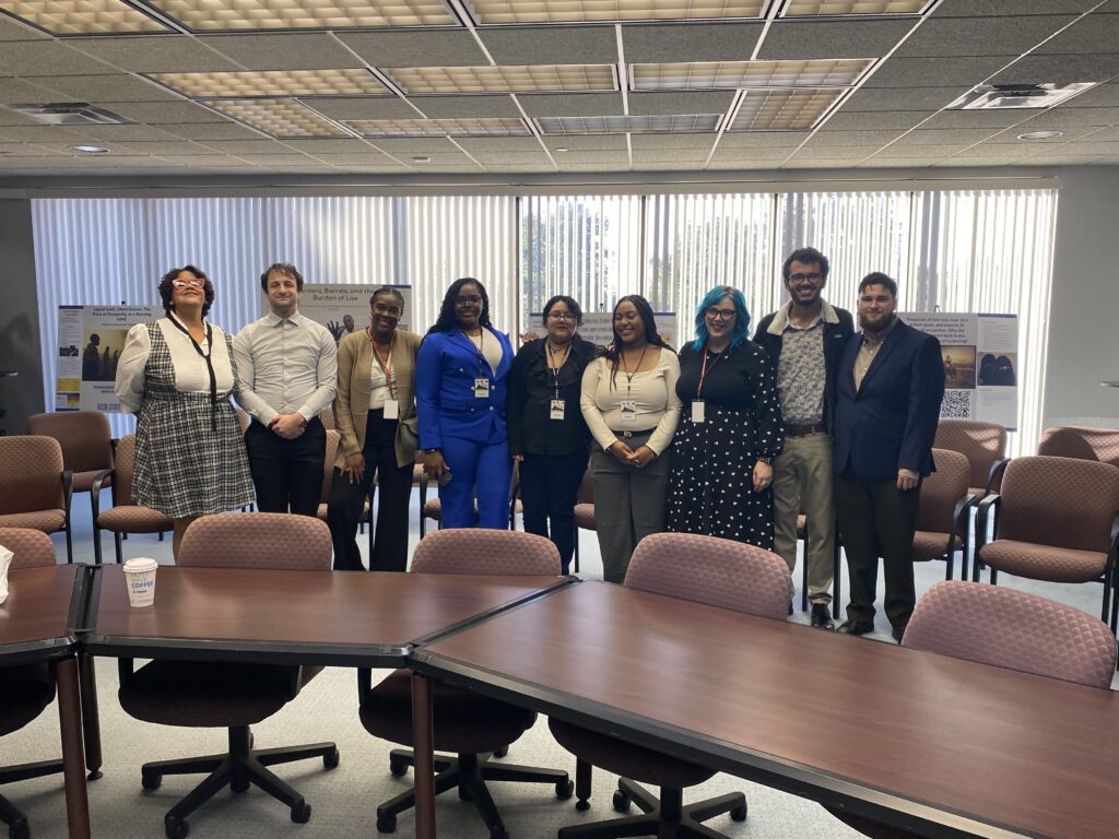 Nine students and faculty members stand together in a conference room with presentation posters displayed behind them, posing for a group photo in front of rows of chairs and tables.