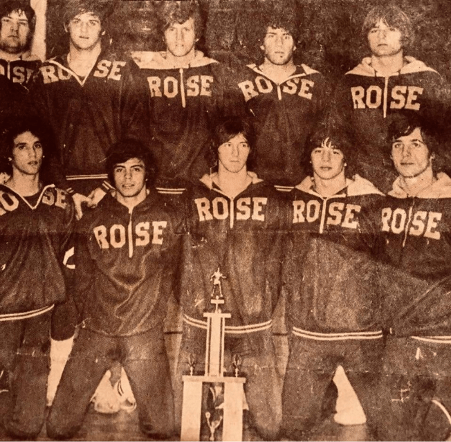 Vintage black-and-white team photo of Rose basketball players wearing “ROSE” warm-up jackets, posed in two rows with a trophy displayed in front.