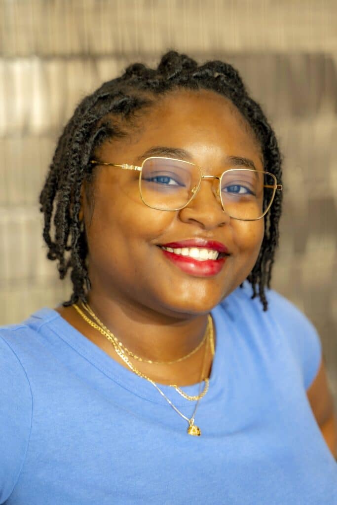 Professional headshot of a smiling woman with short braided hair wearing gold glasses, a blue top, and gold necklaces, in front of a softly blurred neutral background.