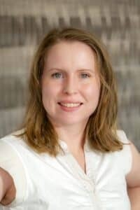 Professional headshot of a smiling woman with shoulder-length light brown hair, wearing a white sleeveless blouse, in front of a softly blurred neutral background.