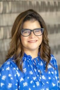 Professional headshot of a smiling woman with long brown hair and glasses wearing a royal blue floral blouse, in front of a softly blurred neutral background.