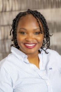 Professional headshot of a smiling woman with short twisted hair wearing hoop earrings and a light blue striped button-down shirt, in front of a softly blurred neutral background.