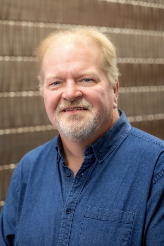 Portrait of a man with short light hair and a beard wearing a blue button-up shirt, smiling in front of a blurred brick wall background.