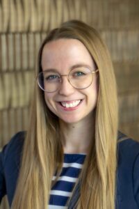Smiling woman with long straight blonde hair and round glasses, wearing a navy blazer over a navy-and-white striped top.