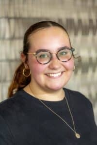 Professional headshot of a smiling woman with glasses and pulled-back hair wearing a black top and gold necklace, in front of a softly blurred neutral background.